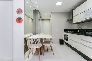 A modern kitchen with white cabinets, a black countertop, and a built-in oven. There is a dining area with a white table and chairs. - Complete Studio 3 Minutes From the sea Jrr205 (Florianópolis)