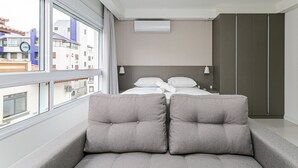 Una habitación de hotel moderna con un ventanal grande, una cama con ropa blanca, un sofá gris con cojines y un armario.