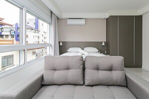 Una habitación de hotel moderna con un ventanal grande, una cama con ropa blanca, un sofá gris con cojines y un armario.