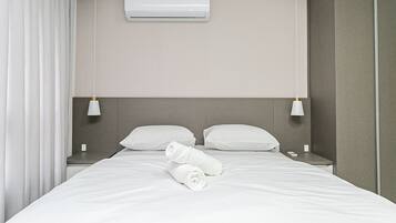 A neatly made bed with white linens and a rolled towel, two white bedside lamps, and an air conditioning unit on the wall.