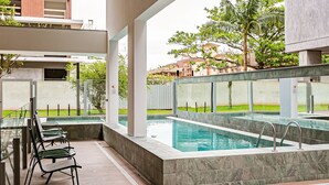 Una piscina con barrera de cristal, mobiliario exterior y vistas a edificios circundantes.