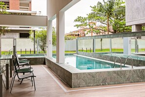 Una piscina con barrera de cristal, mobiliario exterior y vistas a edificios circundantes.