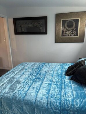 A bedroom with a blue quilted bedspread, a framed picture above the bed, and a door to the left.