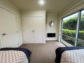 A bedroom with two beds, a white wardrobe, a mirror, and a window with a view of greenery. - Sunny Wānaka Home with Stunning Mountain Views (Wānaka)
