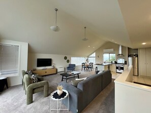 A modern living room with a sofa, a television, and a dining area with chairs and a table. - Sunny Wānaka Home with Stunning Mountain Views (Wānaka)
