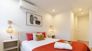 Quarto de hotel com cama, mesas de cabeceira, ar-condicionado fixado na parede, uma obra de arte emoldurada e uma porta.