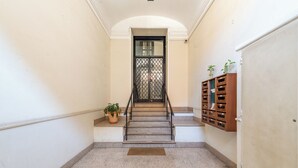Un pasillo con una escalera que lleva a una puerta con celosía metálica. A la izquierda hay una planta en maceta y, a la derecha, un armario de madera con varios compartimentos.