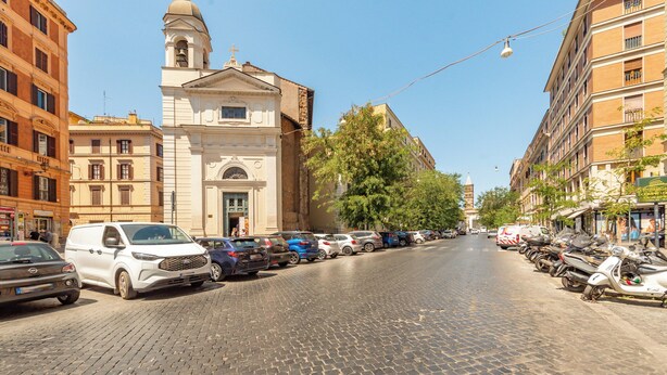 Una calle adoquinada bordeada por vehículos estacionados y edificios, incluyendo una iglesia con campanario.