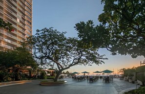 Un patio con mobiliario exterior, un árbol grande y un edificio alto al fondo.