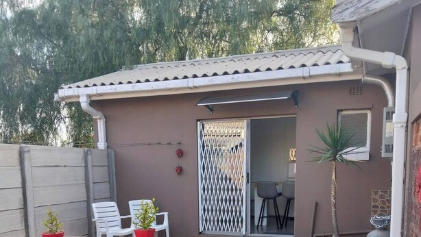 Um pátio com mesa e cadeiras de madeira, uma pequena área de jardim com grama sintética, e um edifício com telhado de telhas e um portão.