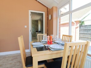 Une table à manger est dressée avec des assiettes, des verres et des salières et poivrières dans une pièce dotée d’une grande fenêtre et offrant une vue sur un bâtiment en briques.