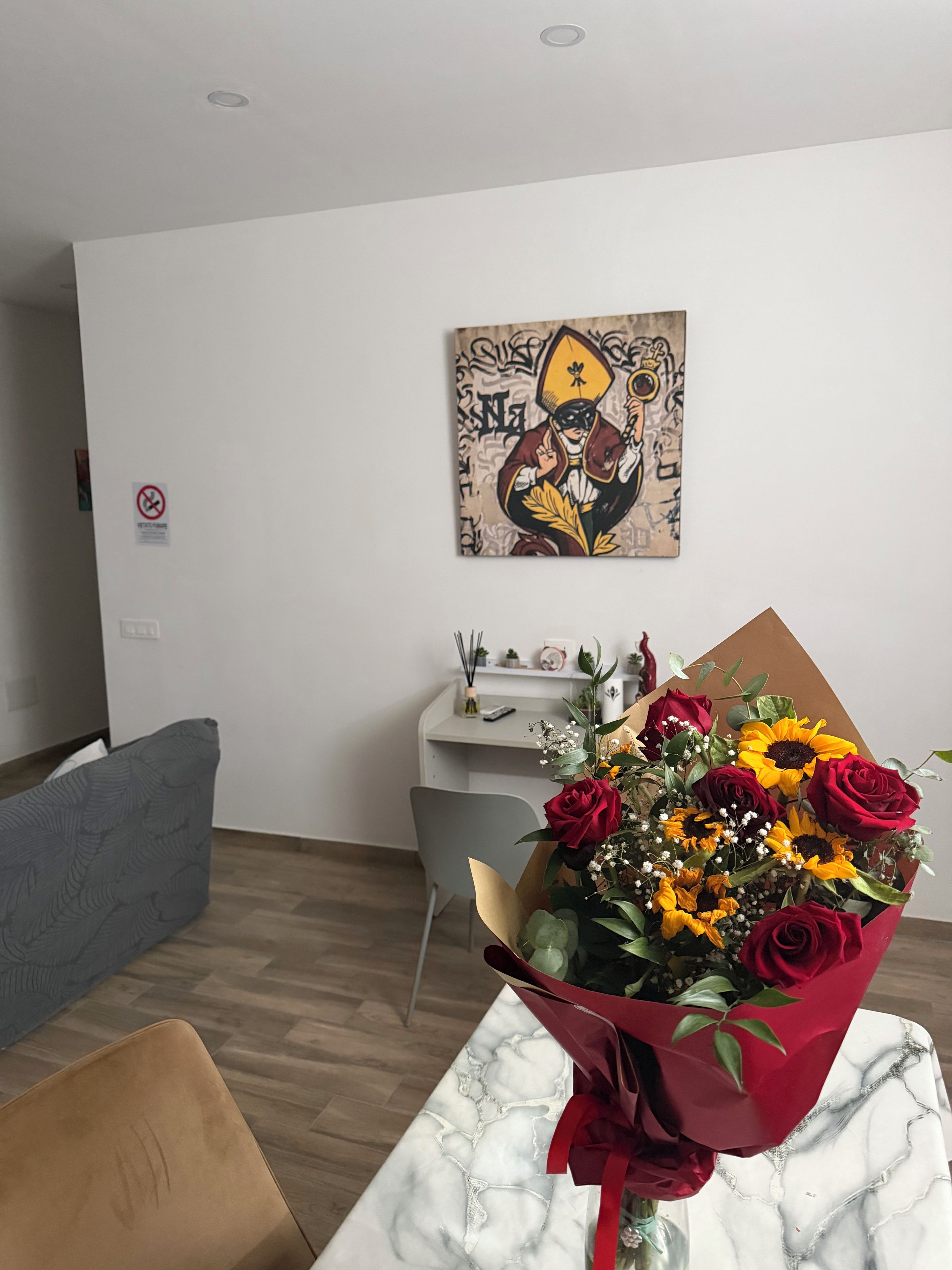 A room with a marble table, a bouquet of flowers, a grey sofa, a white desk, and a wall with a piece of abstract art.
