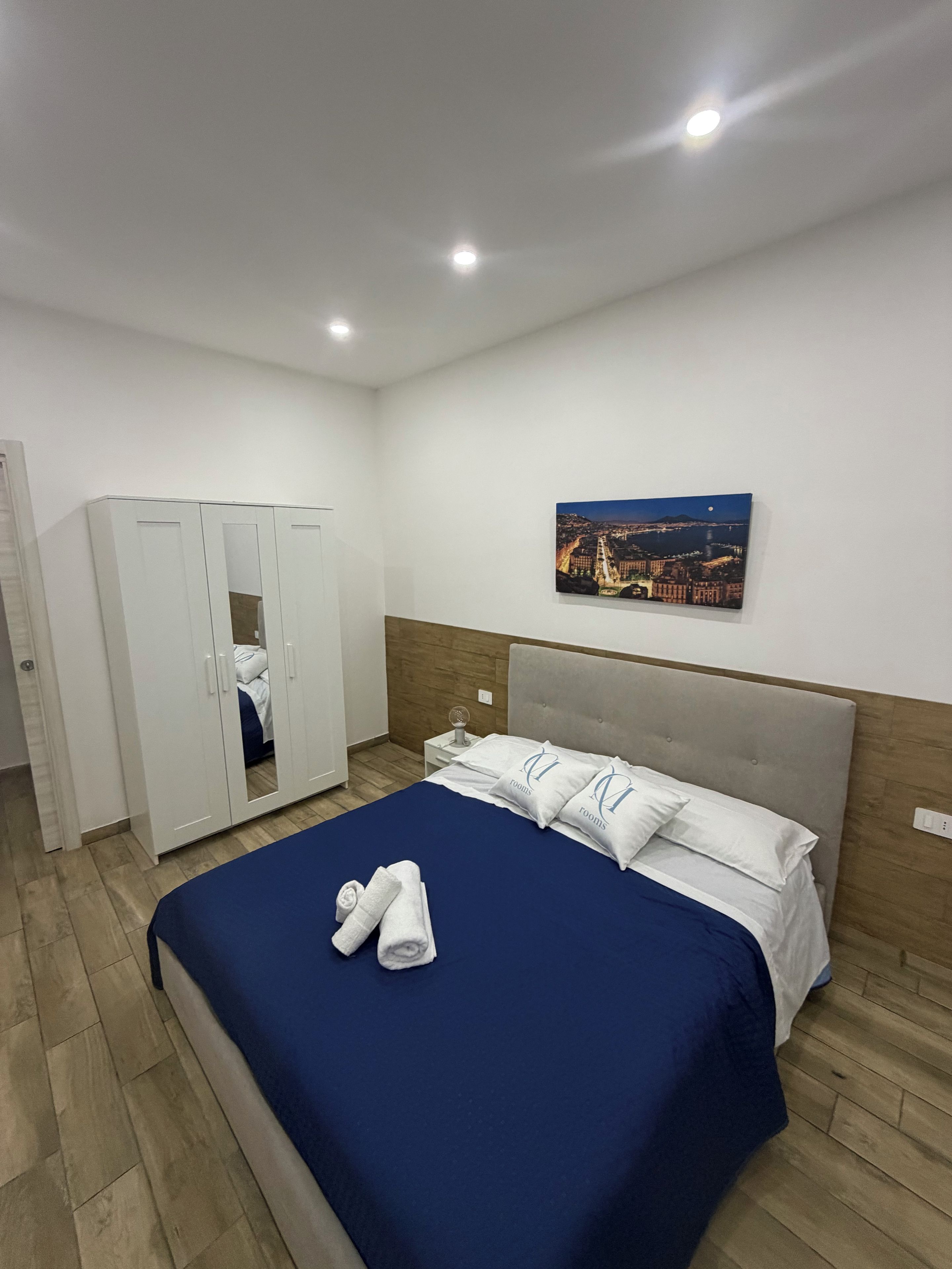 A hotel room with a bed, a wardrobe, a nightstand, a wall-mounted painting, and a wooden floor.