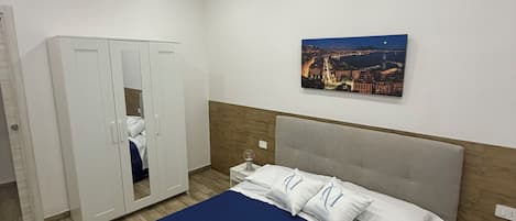 A hotel room with a bed, a wardrobe, a nightstand, a wall-mounted painting, and a wooden floor.
