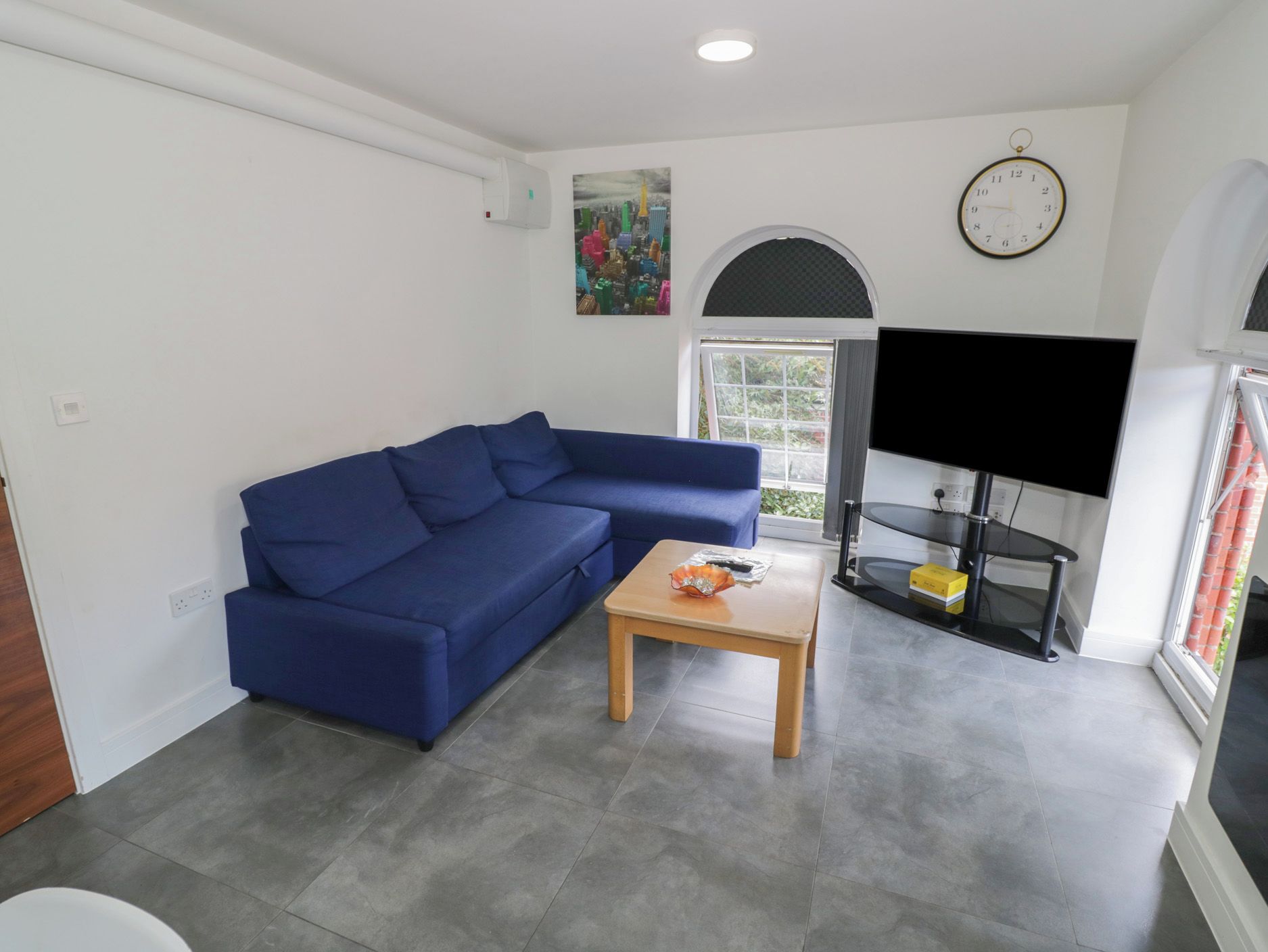 Ein Wohnzimmer mit einem blauen Sofa, einem kleinen Holztisch, einem Flachbildfernseher und einer Uhr an der Wand.