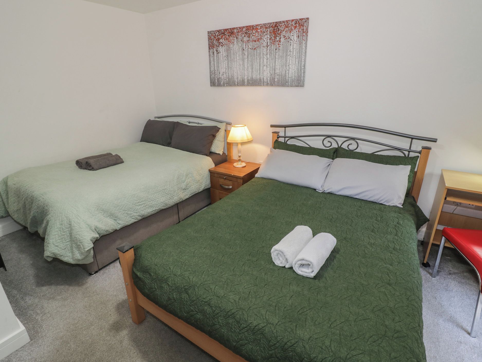 Cama doble con ropa de cama verde y dos toallas enrolladas, una mesita de noche con lámpara, y un escritorio pequeño con una silla.