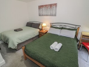 Cama doble con ropa de cama verde y dos toallas enrolladas, una mesita de noche con lámpara, y un escritorio pequeño con una silla.