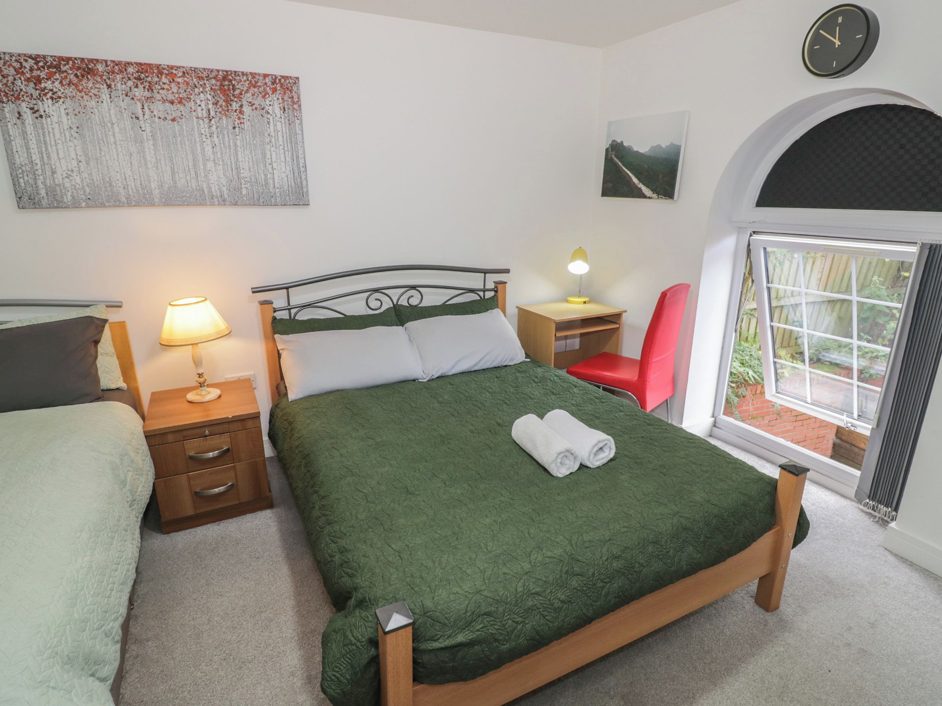 Ein Schlafzimmer mit Bett, Nachttischen, Schreibtisch, Stuhl, einer Uhr und einem Fenster.