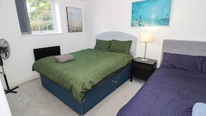 A bedroom with a bed, a bedside table, a lamp, a fan, and artwork on the wall.