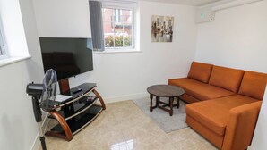 A living room with a flat-screen TV on a wooden stand, an orange sofa, a small round table, and a window with a view of buildings.