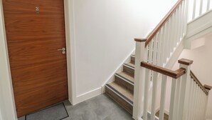 A wooden door with a number plate, a staircase with white railings and wooden handrails, and a grey carpeted floor.
