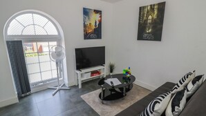 A modern living room with a flat-screen TV, a coffee table, and a sofa with striped cushions. There are two posters on the wall, and a large window with a view of buildings outside.