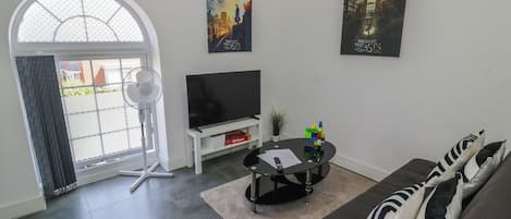 A modern living room with a flat-screen TV, a coffee table, and a sofa with striped cushions. There are two posters on the wall, and a large window with a view of buildings outside.