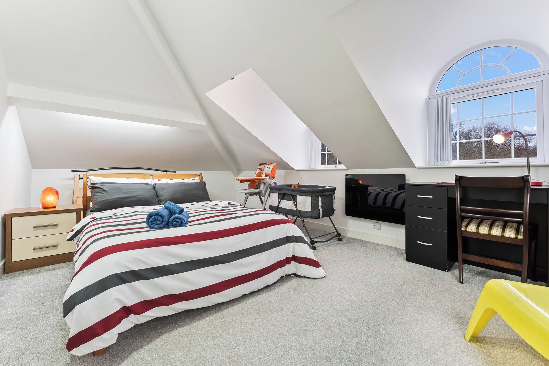 Un dormitorio con techo inclinado, cama con ropa de cama a rayas, escritorio, silla y una mesita de noche con una lámpara.
