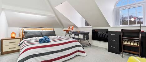 A bedroom with a sloped ceiling, a bed with striped bedding, a desk, a chair, and a small table with a lamp.