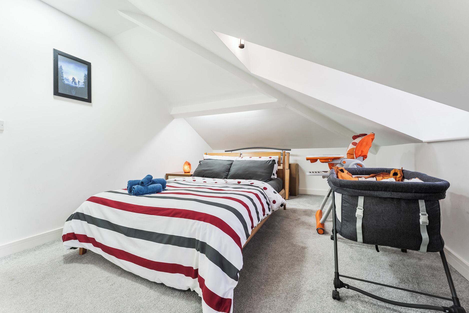 A bedroom with a sloped ceiling, a bed with striped bedding, a nightstand with a lamp, and a baby stroller.