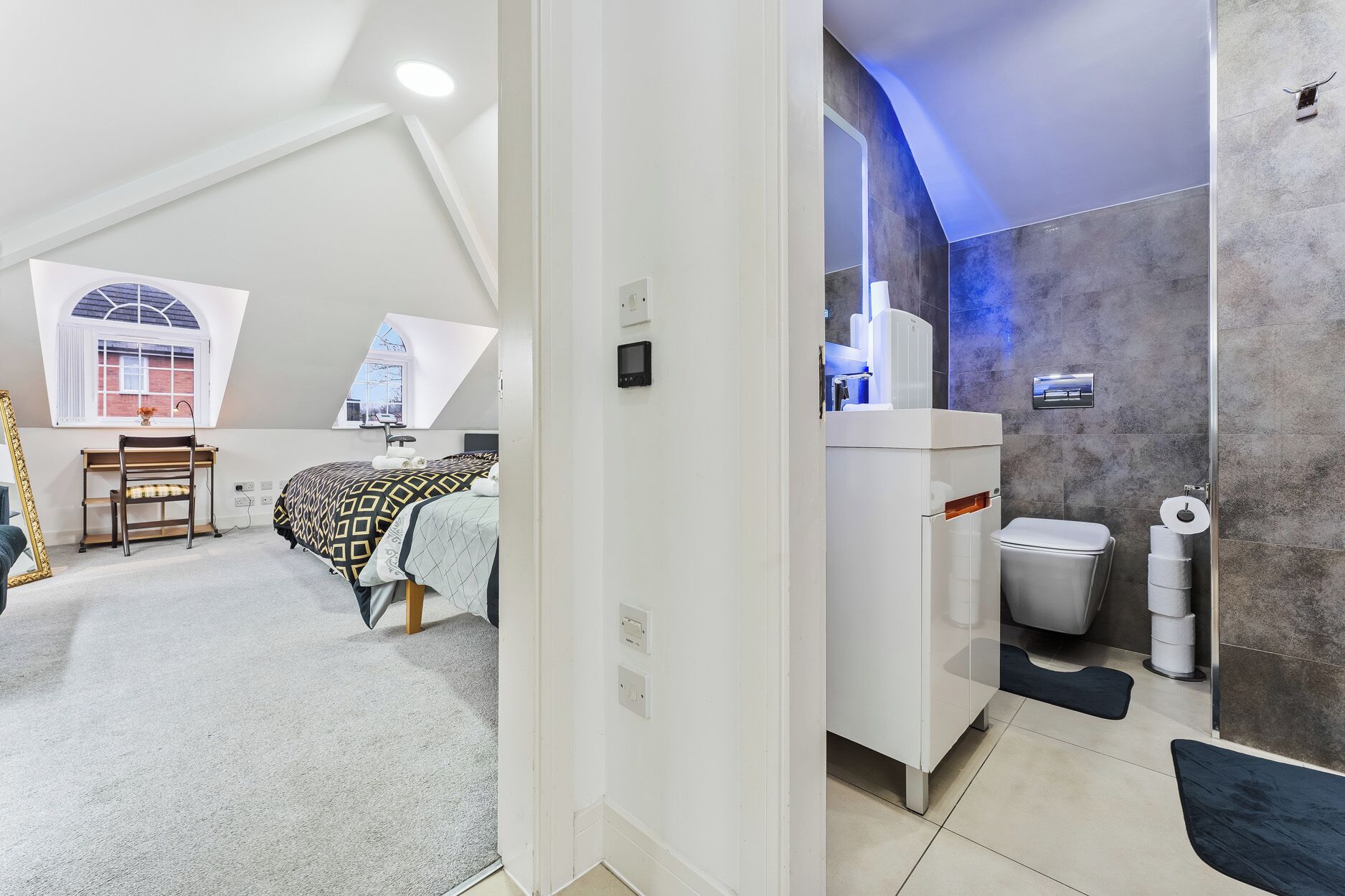 A bedroom with a sloped ceiling, a desk, a bed with patterned bedding, and a window.