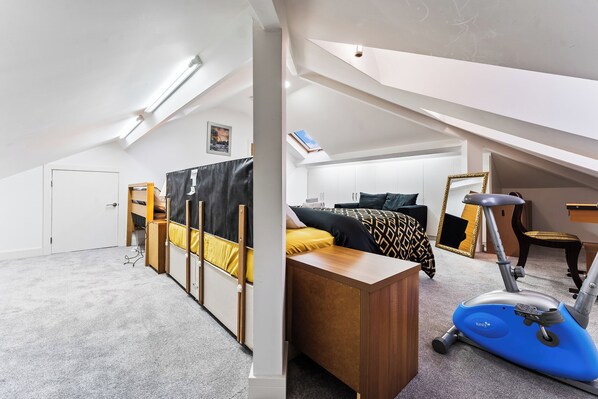 Une petite chambre mansardée, bien éclairée, avec un lit superposé, un bureau, une chaise et un vélo d’appartement bleu.