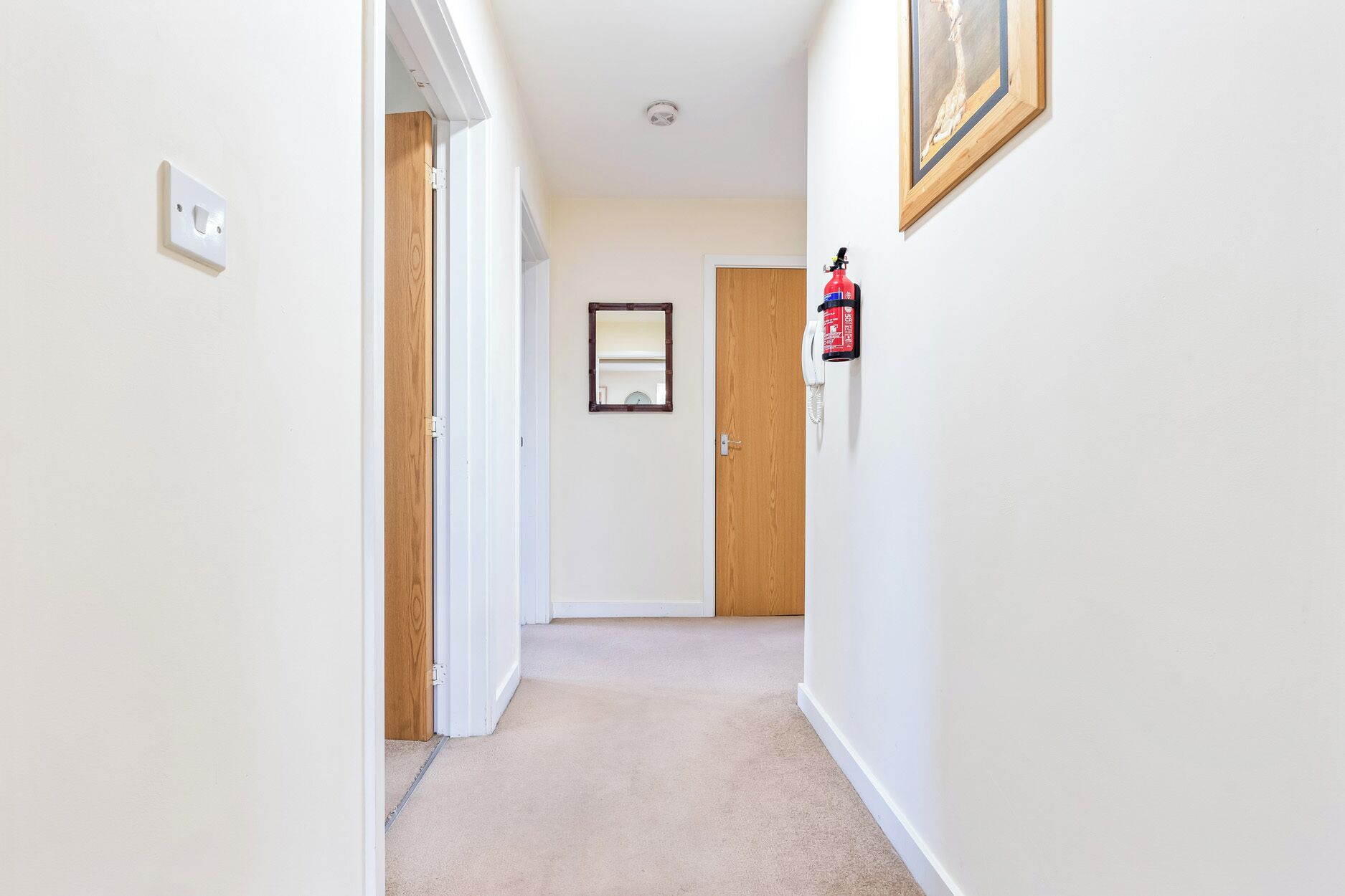 A hallway with carpeted floors, wooden doors, a white wall, and a fire extinguisher mounted on the wall.