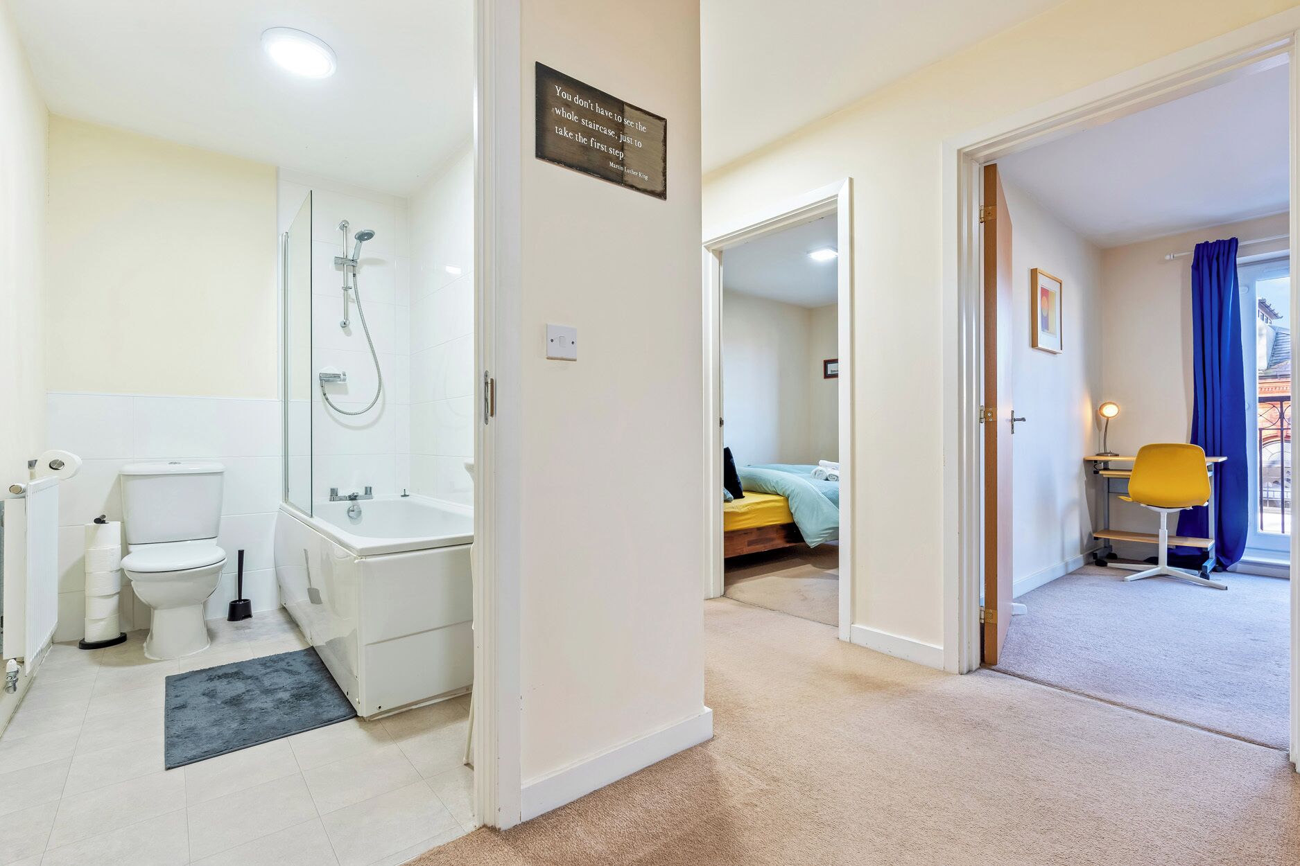 Un baño de hotel con inodoro, lavabo y ducha. Hay un cartel en la pared y una puerta que da a un dormitorio.