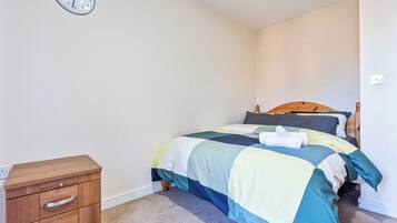 A bedroom with a bed, a wooden nightstand, a wall clock, and a carpeted floor.