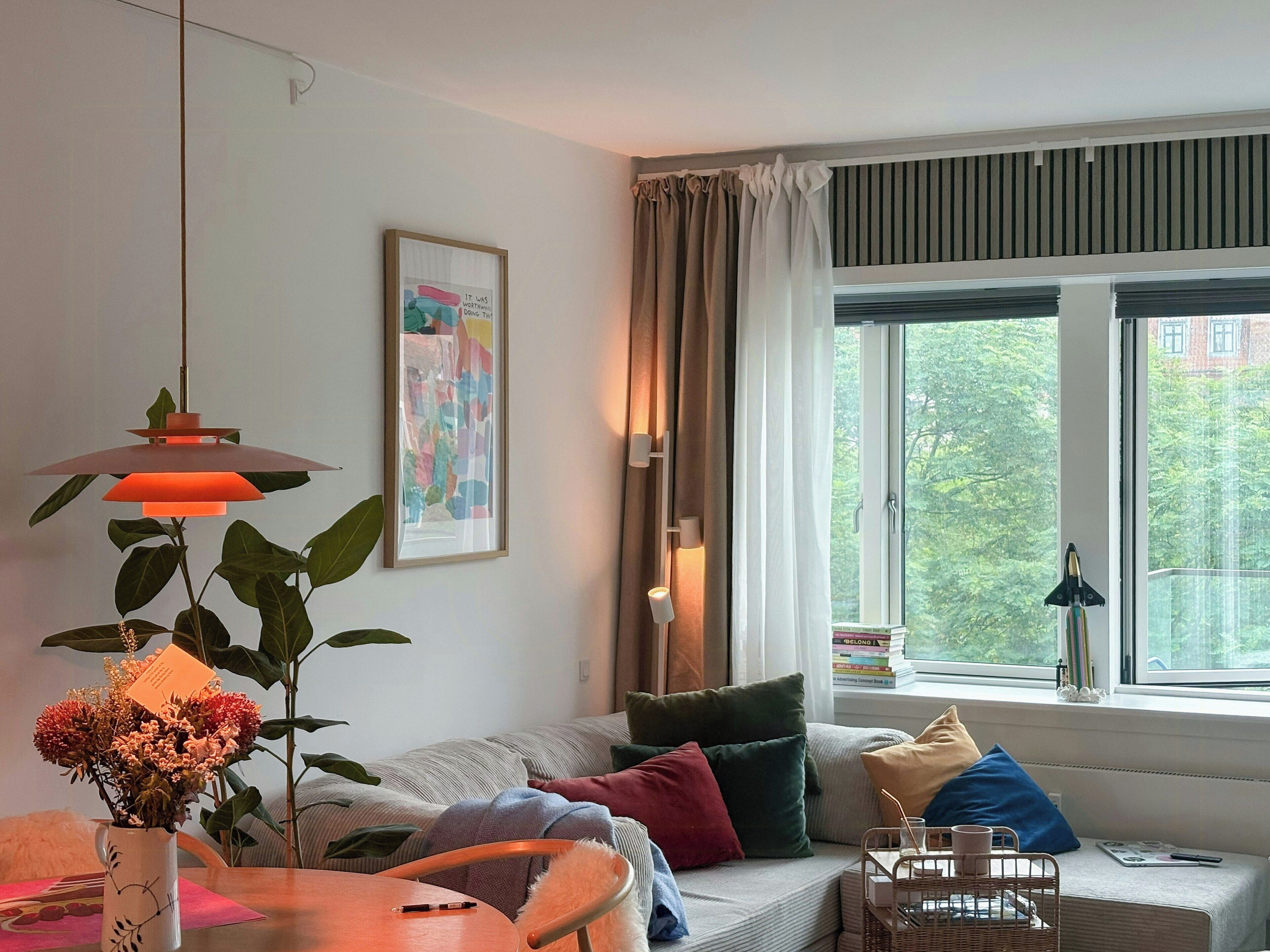 A living room with a sofa, a table with a vase of flowers, a hanging lamp, a framed picture on the wall, and a window with a view of trees.