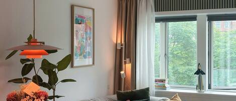 A living room with a sofa, a table with a vase of flowers, a hanging lamp, a framed picture on the wall, and a window with a view of trees.