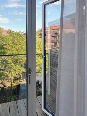 A view from a balcony with a glass door, overlooking a wooden deck and a tree-lined street. - Light flat on Islands Brygge with balcony (København)
