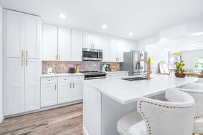 A modern kitchen with white cabinetry, a central island with bar stools, and stainless steel appliances. - The Salty Siren's Cove 3BDR (Tampa)