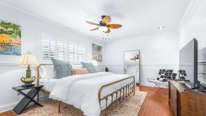 Un dormitorio con cama, mesita de noche, lámpara, ventilador de techo, cómoda, espejo y obras de arte en las paredes.