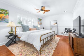 Ein Schlafzimmer mit einem Bett, einem Nachttisch, einer Lampe, einem Deckenventilator, einer Kommode, einem Spiegel und Kunstwerken an den Wänden.