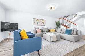 Een moderne woonkamer met een hoekbank, een blauwe fauteuil, een flatscreen televisie en een trap met houten leuningen.