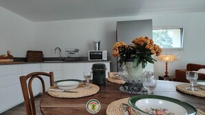 A modern kitchen with white cabinets, a microwave, and a sink. There's a dining table with a vase of flowers and various dishes. - La Grange d'Evelyne 4**** (DIZIMIEU)