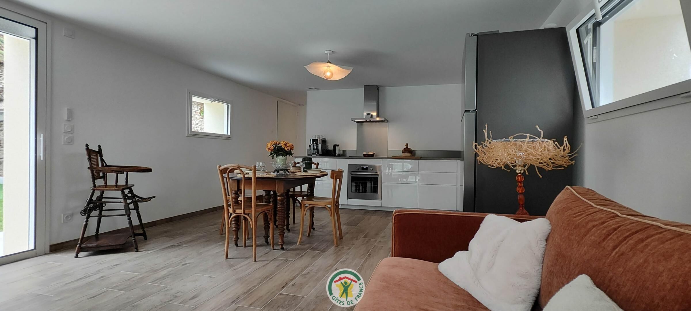 A modern living room with a sofa, dining table, and chairs, a kitchen area with appliances, and a large window providing natural light.