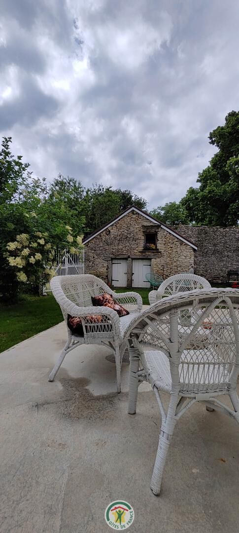 Un bâtiment en pierre avec un toit à deux versants, deux chaises en osier blanches dotées de coussins et une terrasse en béton.