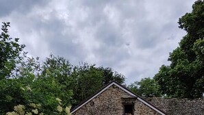 A stone building with a pitched roof, two white wicker chairs with cushions, and a concrete patio. - La Grange d'Evelyne 4**** (DIZIMIEU)
