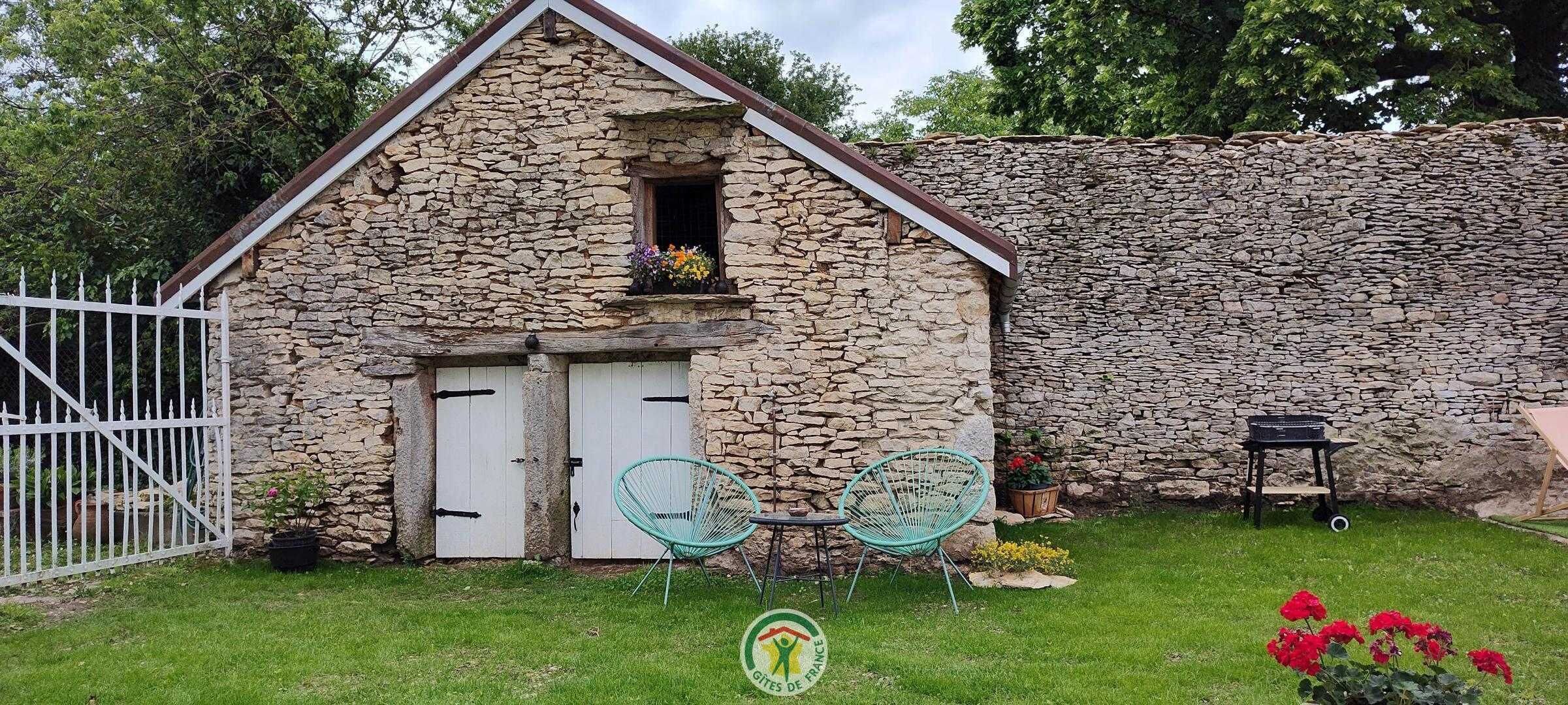 A stone building with a white door and window, surrounded by a white fence, and two green chairs in the foreground.