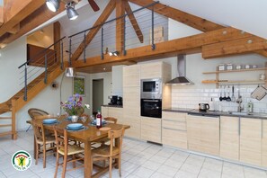 Une cuisine moderne avec des poutres en bois, un coin repas avec une table dressée pour quatre personnes, et un escalier menant à un niveau supérieur.