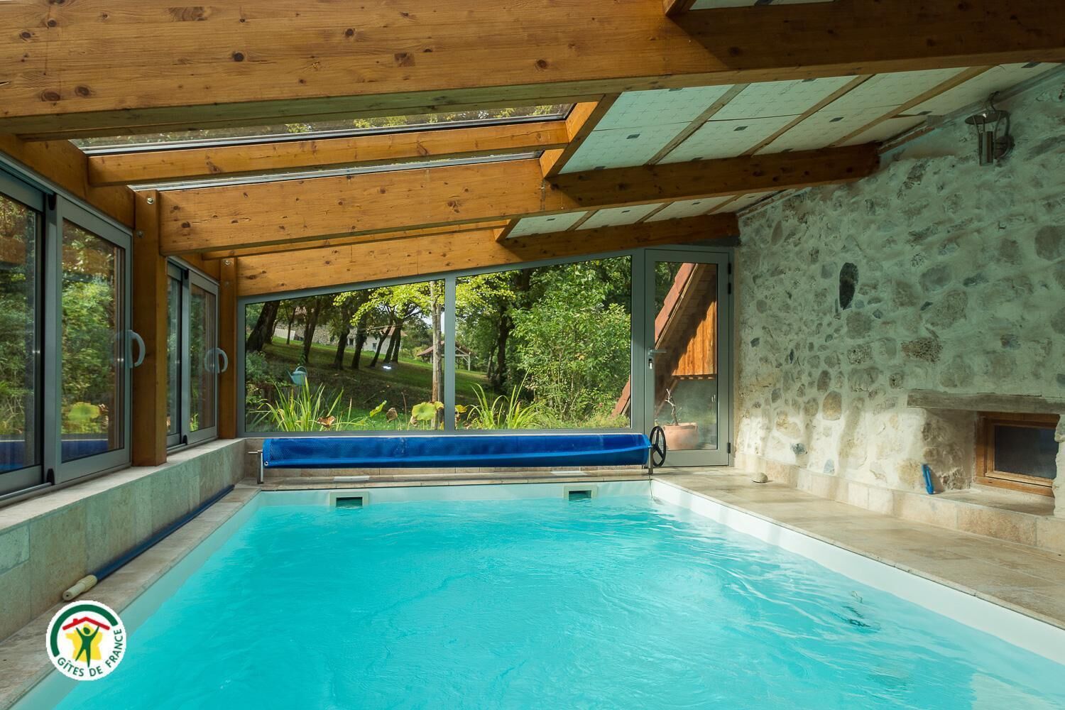 Une piscine dotée d’un plafond en bois et de murs en pierre, de grandes fenêtres et offrant une vue sur la verdure.