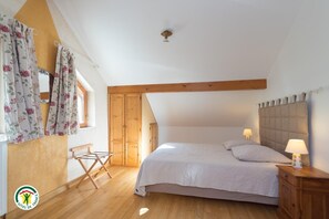 Ein Schlafzimmer mit schrägen Wänden, Holzbalken, einem Bett mit Kissen, einem Nachttisch mit Lampe, einem Holzstuhl und einem Fenster mit Vorhängen.